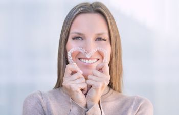 Woman with perfect smile holding invisible dental aligners put in shape of heart in front of her teeth.